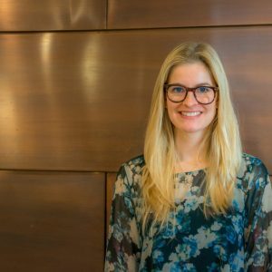 Smiling woman with glasses and long blonde hair, wearing a floral top, stands against a wooden background.