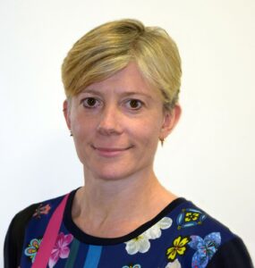 A person with short blonde hair smiles, wearing a colorful floral top on a plain background.