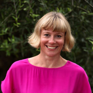 Smiling person with short blonde hair wearing a bright pink top, standing in front of greenery.