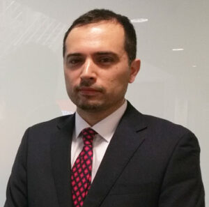 A man in a black suit and red tie stands against a light background, looking at the camera.