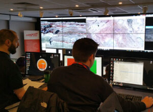 People working at computer workstations in a control room with large screens displaying maps and data.