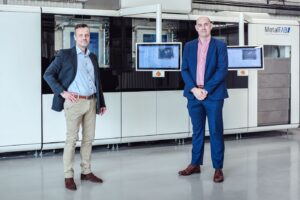 Two men in business attire stand in front of a large industrial metal 3D printer labeled MetalFAB.
