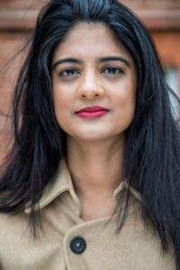 Person with long dark hair and red lipstick wearing a beige coat, standing in front of a brick wall.