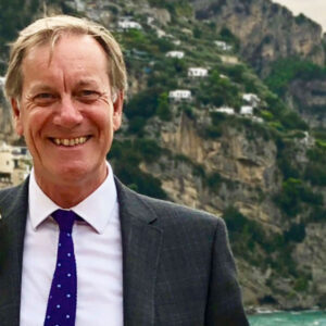 Smiling man in a suit with a polka dot tie stands in front of a scenic cliffside with buildings and greenery.