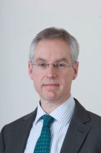 Man with glasses wearing a suit, white shirt, and teal tie, standing against a plain white background.