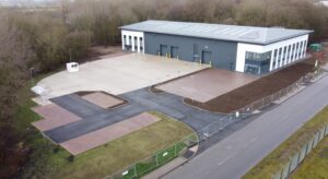 Industrial building with loading bays, surrounded by a paved area, fenced lot, and adjacent road in a wooded area.