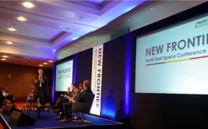 People on stage at the "New Frontier: North East Space Conference" with large screen and audience.