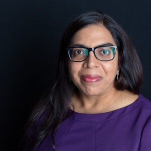Smiling person with long dark hair and glasses, wearing a purple top against a dark background.