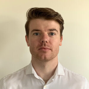 Man with short brown hair and light stubble, wearing a white shirt, against a plain background.