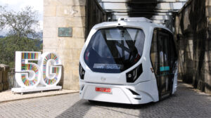 Futuristic white 5G shuttle bus parked beside a colorful "5G" sign and stone wall.