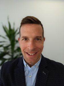 A smiling person in a suit and blue shirt stands indoors with a plant in the background.