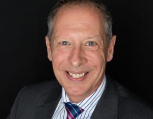 A smiling man in a suit and striped shirt with a red and blue tie against a dark background.