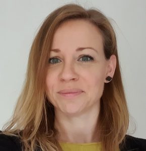 Woman with shoulder-length blonde hair and blue eyes, wearing a green top and black jacket, smiles at the camera.