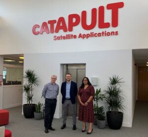 Three people standing in an office lobby under a "CATAPULT Satellite Applications" sign, surrounded by plants.
