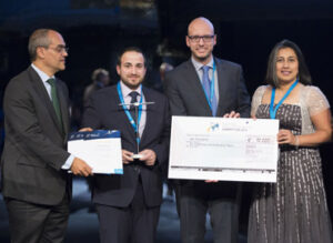 Four people in formal attire are standing, holding a large ceremonial check and certificates on stage.