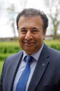 Smiling man in a suit and tie standing outdoors, with trees blurred in the background.