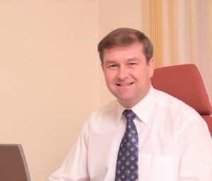 Smiling man in a tie sits at a desk with a laptop and papers in an office setting.