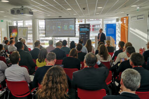 A speaker presents to a seated audience in a conference room with large screens and banners.
