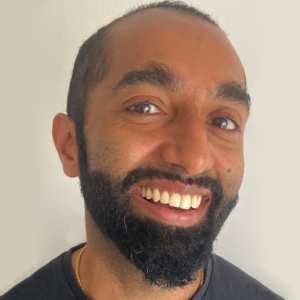 Smiling man with a beard, wearing a black shirt and gold necklace, against a plain background.