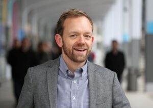 Man with a beard wearing a gray blazer, standing outdoors, smiling with a blurred background of people walking.