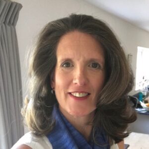 Woman with long brown hair and a blue scarf, smiling indoors near a light gray curtain.