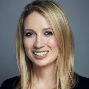 Smiling woman with long blonde hair wearing a black top, against a gray background.