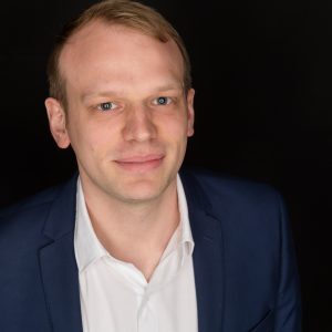 Man in a blue suit with a white shirt, smiling against a black background.