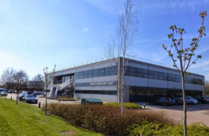 Modern two-story office building with parked cars and surrounding greenery on a sunny day.