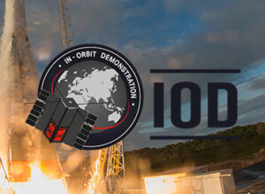 Graphic of a satellite labeled "In-Orbit Demonstration" with "IOD" text, set against a cloudy sky and rocket launch.