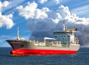 A large cargo ship with a red and white hull navigates through calm ocean waters under a cloudy sky.