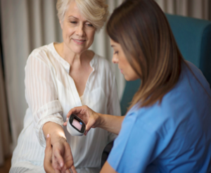 A nurse checks an older woman's blood pressure with a digital monitor. They are seated in a light-filled room.