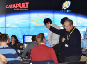 People collaborating in a tech room with screens displaying earth and logos, including "CATAPULT" and "TESTFEST.