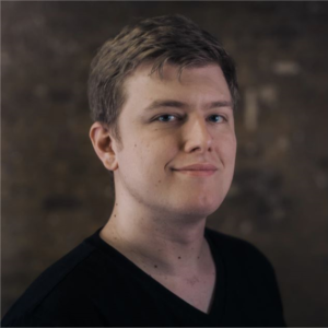 Person with short hair wearing a black shirt, smiling slightly against a blurred brown background.