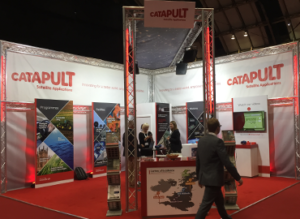 Exhibition booth with red elements and "CATAPULT" branding, displaying satellite applications.