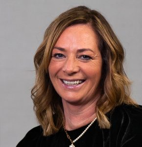 Smiling woman with shoulder-length wavy hair wearing a black top and a pendant necklace, against a plain background.