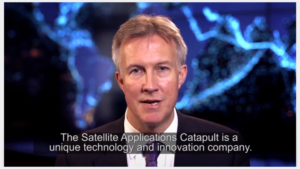 A man in a suit speaks in a video with a world map background. Subtitles mention Satellite Applications Catapult.