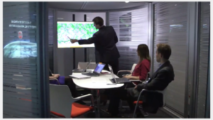 A person presenting data on a large screen to three people seated around a table in a modern office.