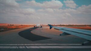 Airplane wing view on runway with another plane ahead under a partly cloudy sky.