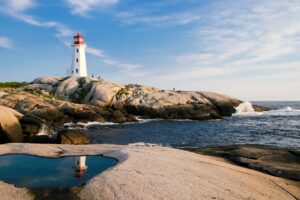 Lighthouse on rocky shore by the ocean with a reflection in a tidal pool, under a blue sky with clouds.