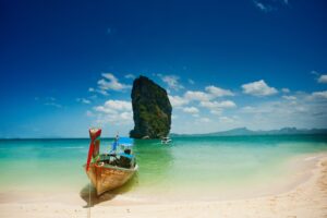 Wooden boat on a sandy beach with turquoise water, clear skies, and a large rock formation in the background.