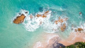 Aerial view of turquoise ocean waves crashing on a sandy beach with scattered rocks.