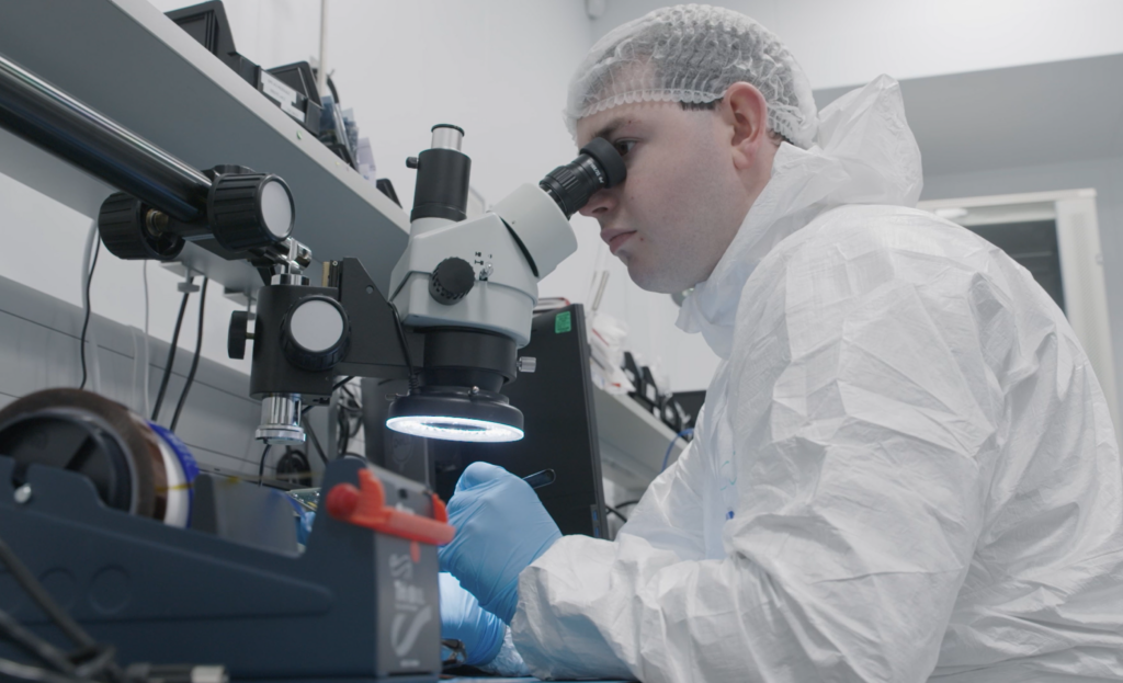 Lab technician in protective gear examines a sample under a microscope in a laboratory setting.