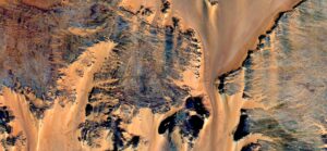 Satellite view of a desert landscape, featuring sand dunes and rocky formations in contrasting patterns and colors.