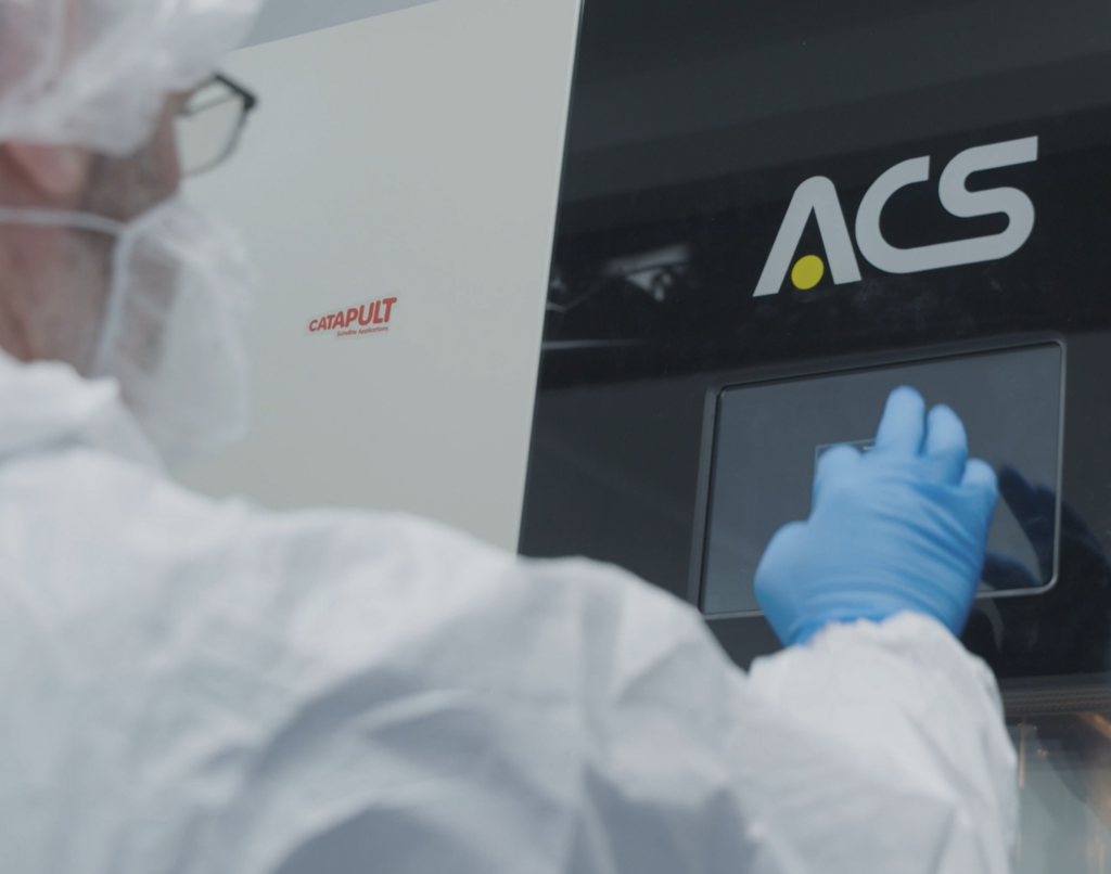 Person in lab gear operating equipment with ACS logo, wearing blue gloves and a protective suit.
