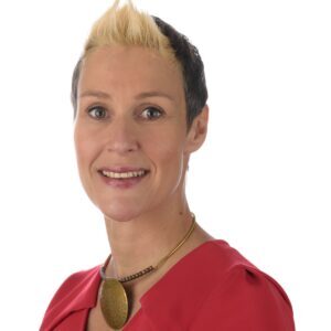 Person with short hair wearing a red shirt and a necklace, smiling against a white background.