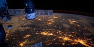 Satellite view of Earth at night showing city lights sprawling across a continent, with part of a spacecraft visible.