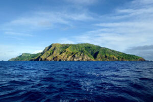 Lush green island with rocky cliffs surrounded by deep blue ocean under a cloudy sky.