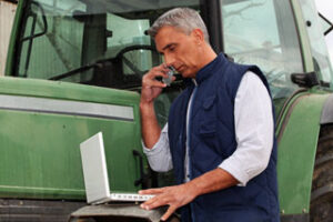 Man in blue vest on phone, using a laptop, standing beside a green tractor.
