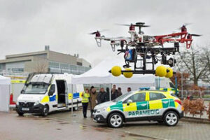 A drone hovers near a paramedic car and a van with people standing nearby in an outdoor setting.