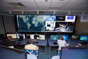 People in a control room with multiple monitors displaying world maps and satellite images.